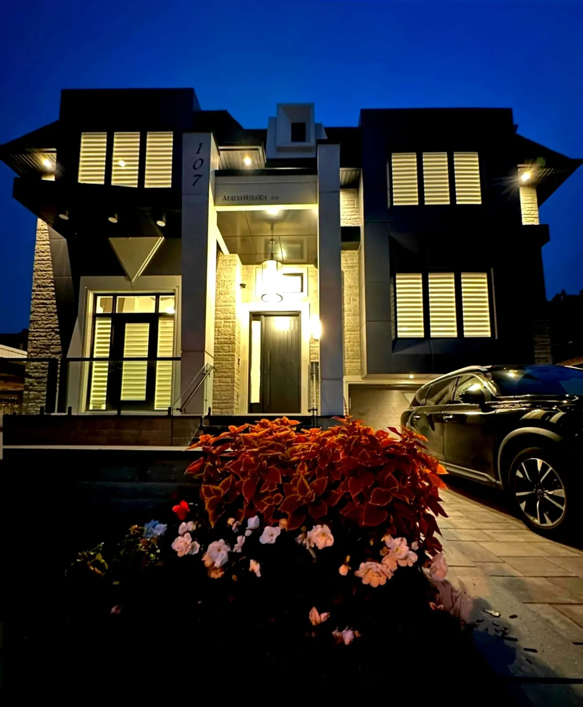 Two-storey house at dusk with black cladding, stone piers and warm lit windows