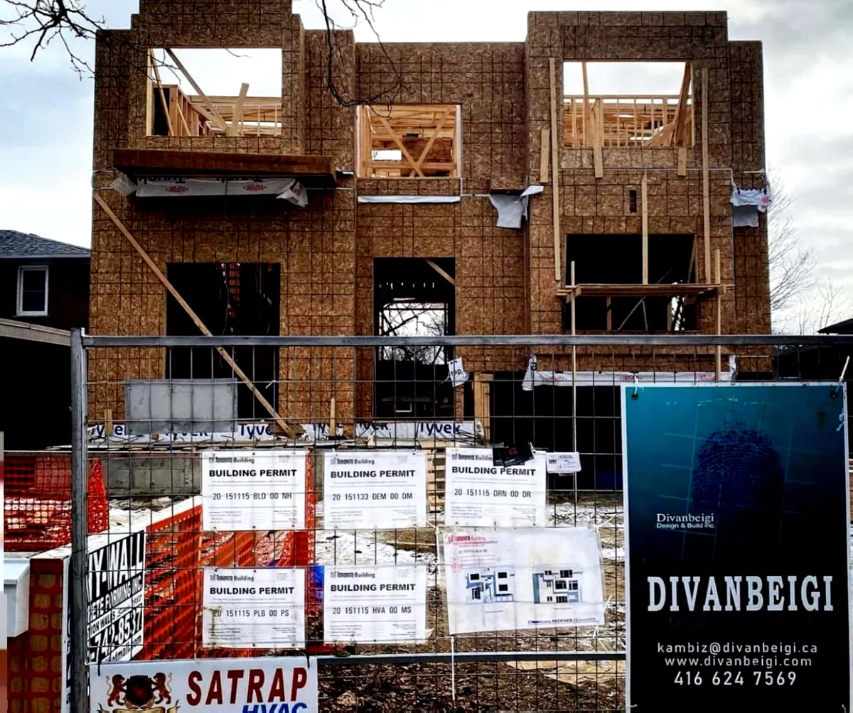 Wood-framed house wrapped in sheathing behind a hoarding with building permits and the Divanbeigi Design & Build sign