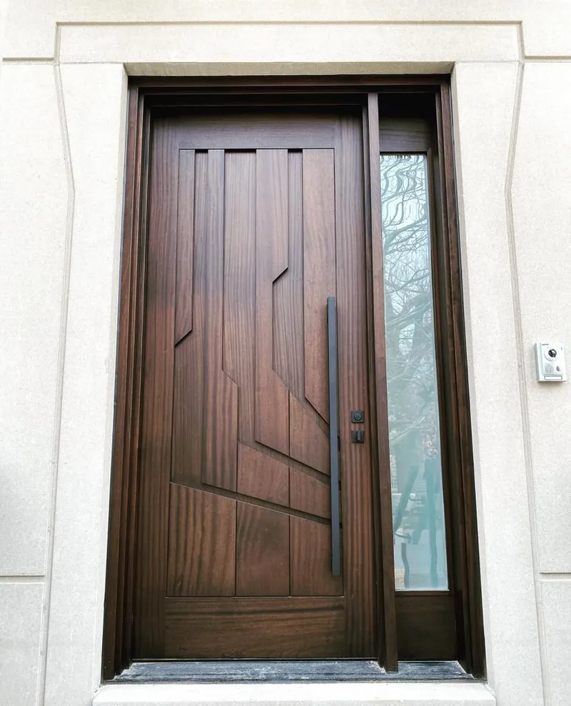 Walnut front door with inlaid geometric panels and a long black handle in a limestone frame